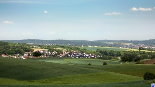Landschaftsaufnahme. Vordergrund: grüne Hügel. Hinten im Bild: Dorf. Der Himmel ist blau.