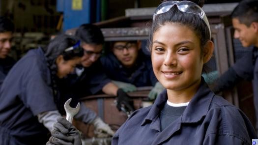 Eine junge nepalesische Frau im Blaumann lächelt in die Kamera. Im Hintergrund arbeiten weitere Jugendliche mit Werkzeugen gemeinsam an einem Projekt.