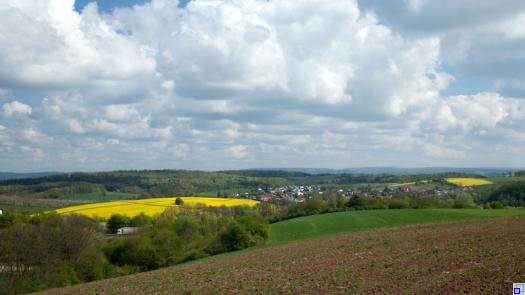 Landschaftsaufnahme mit Hügeln in Braun (leerer Acker), Grün (Feld und Wald) und Gelb (Rapsfeld), darüber ein blauer Himmel mit weißen Wolken