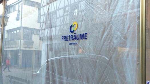 Außenaufnahme eines Schaufensters, das von innen mit Folie behängt ist. Im Fenster spiegeln sich die Fachwerkbauten auf den gegenüberliegenden Straßenseite. Das FREIRÄUME-Logo prangt auf der Scheibe.