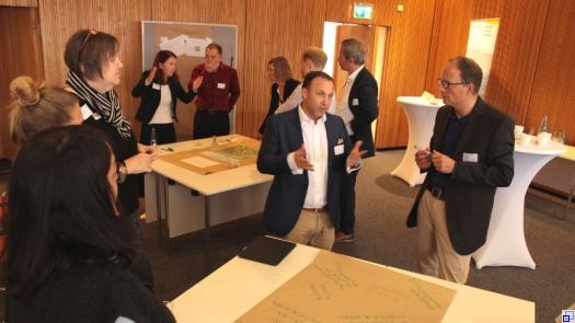Mehrere Menschen diskutieren an einem Tisch, auf dem ein handschritflich beschriebenes Plakat liegt. Im Hintergrund ein weiterer Tisch mit Diskutierenden.