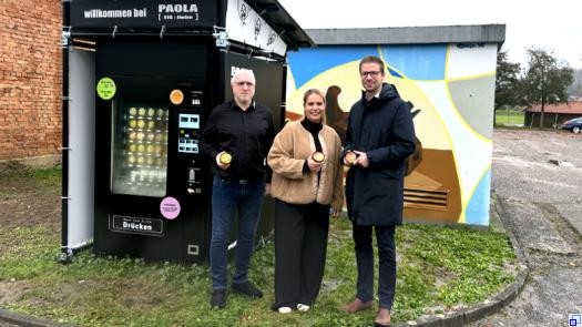 Zwei Männer und eine Frau stehen in Winterjacken vor einem gut gefüllten neuen Eisautomaten und lächeln in die Kamera.