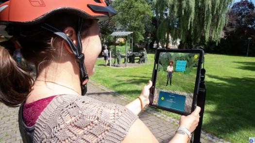 Frau mit Fahrradhelm hält ein Tablet in der Hand, auf dessen Bildschirm eine Frau in der Umgebung zu sehen ist, in der die Fahrradfahrerin steht.