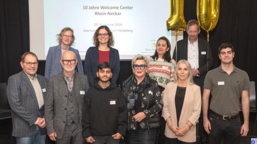 Eine Gruppe Menschen mit eher förmlicher Kleidung steht vor einer Präsentation mit der Aufschrift "10 Jahre Welcome Center Rhein-Neckar"