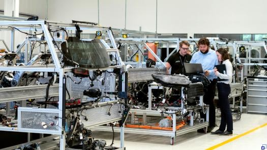 Drei Menschen unterschiedlicher Geschlechter stehen in einer Produktionshalle und blicken gemeinsam auf einen Laptop.