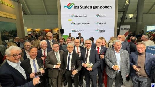 Mehrere Männer im Anzug stehen in einer Messehalle vor einer Werbewand mit dem Slogan "Im Süden ganz oben"