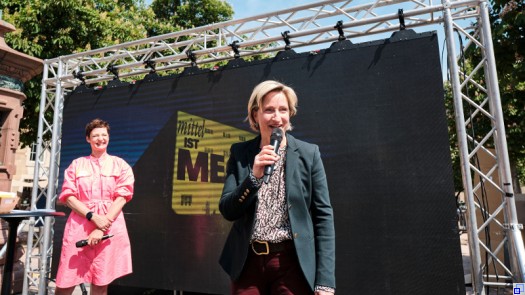Dr. Nicole Hoffmeister-Kraut steht im Business-Kostüm auf einer Bühne, im Hintergrund ist eine weitere Frau sowie ein Banner mit der Aufschrift mittel IST MEGA zu sehen.