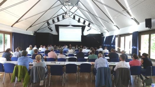 In einer Halle mit hohem weißen Dach sitzen zahlreiche Menschen an langen Tischreihen und blicken auf eine Präsentation ganz vorne im Raum.