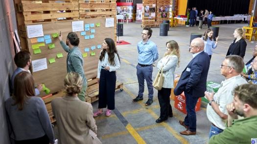 mehrere Menschen in Businesskleidung stehen in einer Halle und betrachten eine improvisierte Pinnwand, an der mehrere bunte Zettel kleben. Ein Mann klebt einen neuen Zettel hinzu.