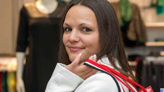 Eine junge Frau mit dunklen Haaren steht vor einem Bekleidungsgeschäft. Sie hat mehrere Einkaufstaschen locker über die Schulter gelegt und lächelt in die Kamera.