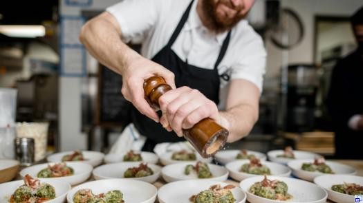 Mann steht in einer Restaurantküche und mahlt aus einer hölzernen Mühle frischen Pfeffer über eine Reihe gleichartiger Vorspeisen..