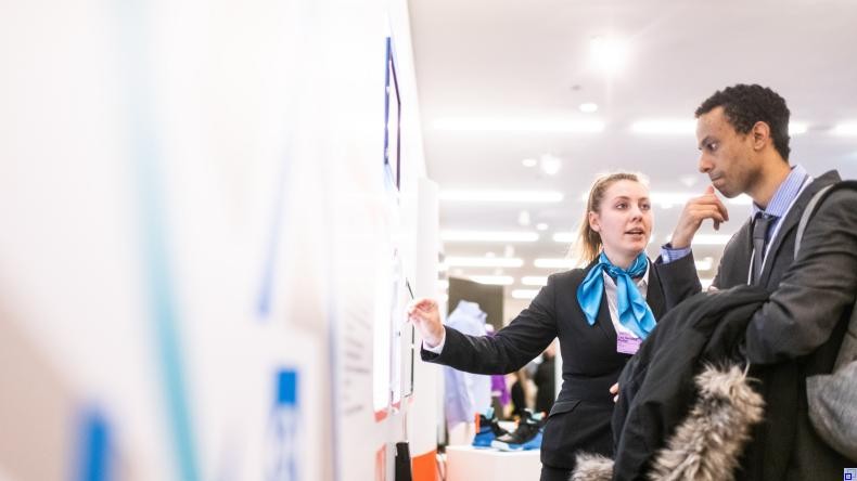 Bild: Evangeline Shaw Junge Frau erklärt jungem Mann etwas. Beide stehen in einer Messehalle und tragen Businesskleidung.