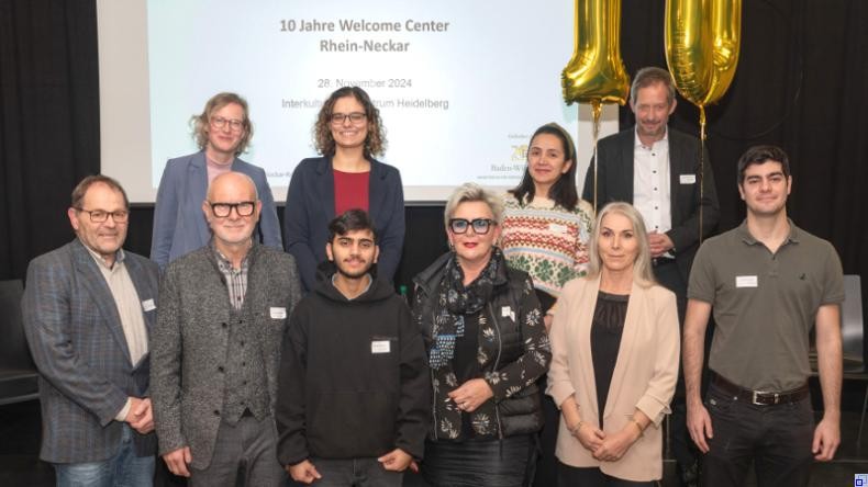 Bild: Burkhardt / Rhein-Neckar-Kreis Eine Gruppe Menschen mit eher förmlicher Kleidung steht vor einer Präsentation mit der Aufschrift "10 Jahre Welcome Center Rhein-Neckar"