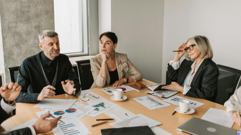 Bild: Vlada Karpovich/Pexels Mehrere Menschen sitzen an einem Konferenztisch, auf dem Unterlagen, Notebooks und Kaffeetassen stehen bzw. liegen. Ein grauhaariger Mann spricht, zwei Frauen mittleren Alters hören zu. Eine blickt skeptisch.