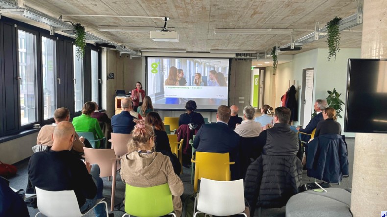 Bild: Stadt Mannheim Ein Raum mit bunten Stühlen; die Menschen, die darauf sitzen, sind von hinten zu sehen. Vor der Gruppe steht eine Frau mit Mikrofon vor einer Bildschirmpräsentation.