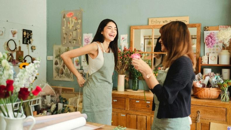 Bild: Amina Filkins / Pexels Junge Frau legt Schürze an und lacht, ältere Frau lächelt ihr zu. Sie stehen in einem Blumenladen hinter der Theke.