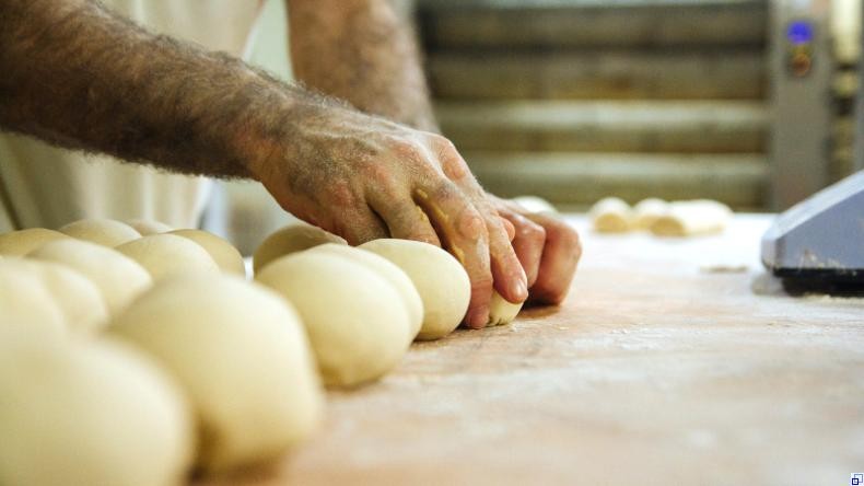 Bild: unsplash.com Eine Reihe Dampfnudeln liegt auf einer bemehlten Arbeitsfläche. Ein Mann, von dem nur die stark behaarten Arme zu sehen sind, formt im Hintergrund Teig zu weiteren Dampfnudeln.