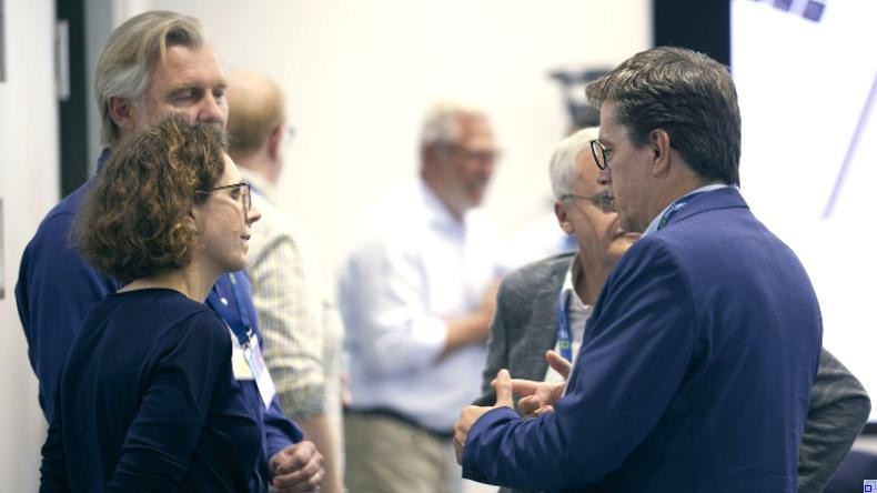 Bild: NSI Eine Vierergruppe von Menschen in Businesskleidung steht beisammen und unterhält sich. Im Hintergrund sind weitere Menschen in Businesskleidung zu sehen.