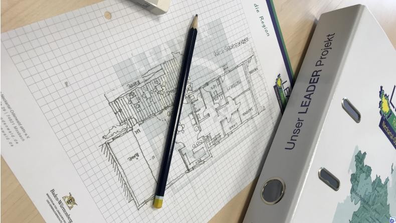 Bild: LEADER Neckartal-Odenwald Symbolbild: Ordner mit der Aufschrift "Unser LEADER Projekt", daneben kariertes Blatt mit gezeichnetem Grundriss, darauf ein Bleistift.