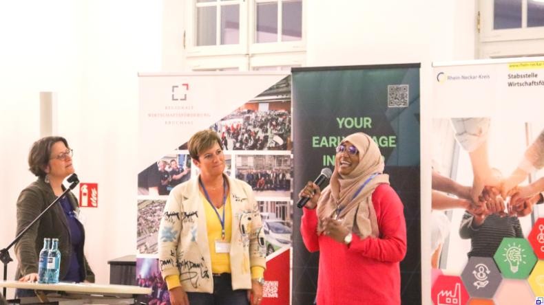 Bild: Kotsch / Rhein-Neckar-Kreis Drei Frauen stehen auf der Bühne; die Dame rechts im roten Kleid und mit Hijab referiert, die anderen beiden hören zu.