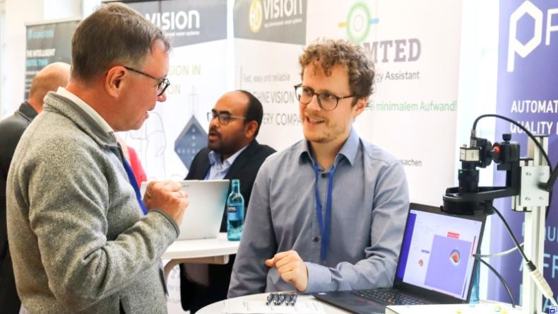 Bild: Kotsch / Rhein-Neckar-Kreis Zwei Männer unterhalten sich an einem Stehtisch, auf dem ein Laptop sowie ein Messgerät aufgebaut ist. Im Hintergrund sind weitere Menschen sowie verschiedene Roll-ups zu sehen.