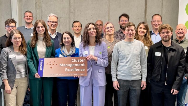 Bild: Stadt Mannheim Eine Gruppe von Menschen steht lächelnd vor einer Betonwand. In der vorderen Reihe stehen drei Frauen in Businesskleidung, die gemeinsam ein großes Plakat mit der Aufschrift "Cluster Management Excellence" halten.