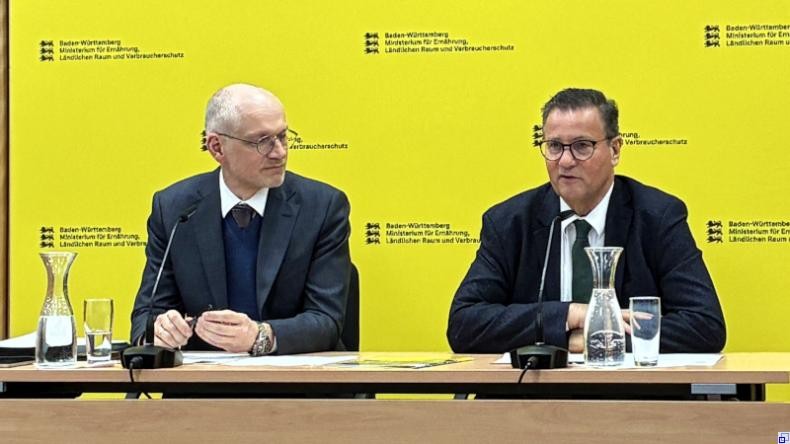 Bild: Ministerium für Ernährung, Ländlichen Raum und Verbraucherschutz Zwei Männer im Businessdress sitzen vor gelbem Hintergrund an einem Schreibtisch mit Unterlagen.