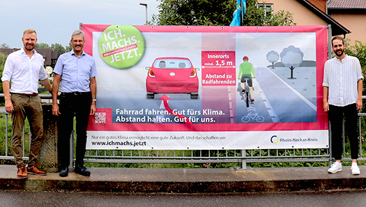 3 Personen stehen vor einem großformatigen Banner mit Informationen zur Abstandsregelung