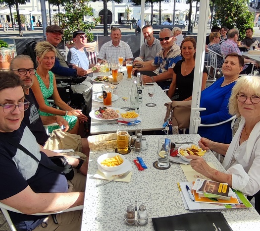 9 Mitglieder des Inklusionsbeirats sitzen lachend an einem Restauranttisch im Freien. Sie essen und trinken.