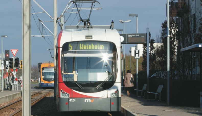 Das Bild zeigt eine Straßenbahn in Frontaufnahme in den Farben weiß-rot an einer Haltestelle. In der digitalen Anzeige steht "Weinheim" und "5" - so heißt die Linie.