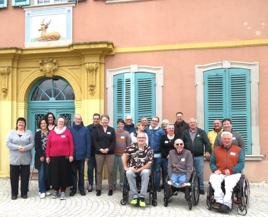 Die 20 teilnehmenden Mitglieder des Beirats stehen als Gruppe vor dem gelben Eingang des Palais Hirsch. Die In ihrer Mitte steht die Behindertenbeauftragte. Über dem Eingang ist ein goldener Hirsch.