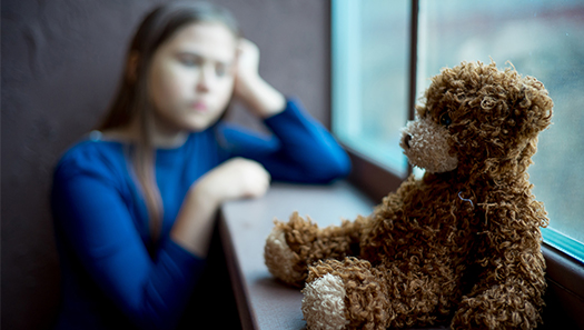 Kind schaut traurig aus dem Fenster, im Vordergrund sitzt ein Teddybär