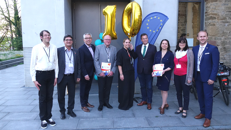 Eine Gruppe von Menschen steht mit einem goldenen "10"-Ballon vor einem EU-Fähnchen.