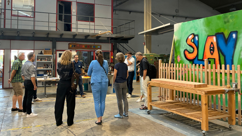 Menschen stehen in einer Werkshalle neben einer Holzkonstruktion und hören einem Mann zu.