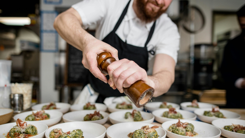 Mann steht in einer Restaurantküche und mahlt aus einer hölzernen Mühle frischen Pfeffer über eine Reihe gleichartiger Vorspeisen..