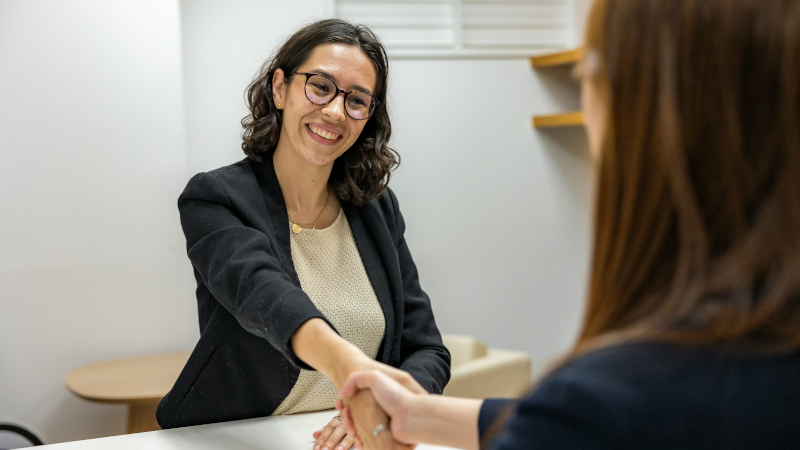Zwei Frauen in Businesskleidung sitzen einander gegenüber, eine davon ist nur angeschnitten von hinten zu sehen. Sie schütteln sich die Hände; die Frau, die von vorne gezeigt wird, lächelt.