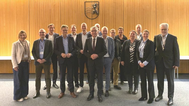Eine Gruppe Menschen, hauptsächlich Männer, steht in einem holzgetäfelten Raum und lächelt in die Kamera. Alle tragen Businesskleidung.