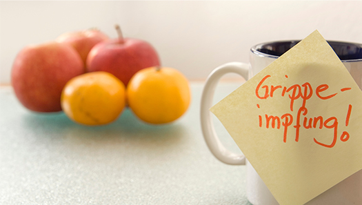 Auf einer Kaffeetasse klebt ein Post-it mit der Aufschrift Grippeimpfung!