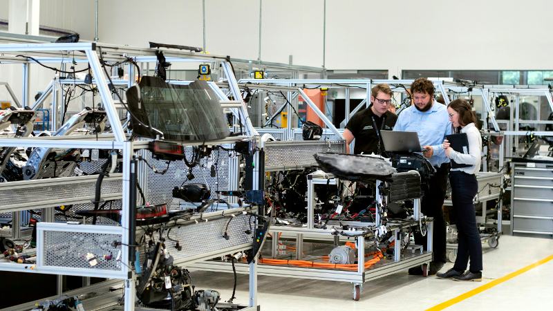 Drei Menschen unterschiedlicher Geschlechter stehen in einer Produktionshalle und blicken gemeinsam auf einen  Laptop.