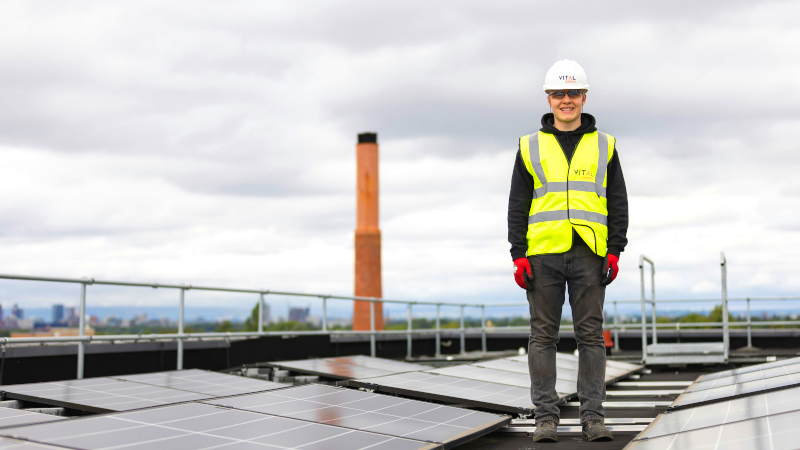 Person in gelber Warnweste steht auf Industriedach zwischen Solarpaneelen.