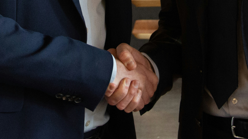 Nahaufnahme: Zwei Männer schütteln sich die Hände. Einer trägt blaues Jackett und keine Krawatte, der andere ein dunkles Jackett und Krawatte.