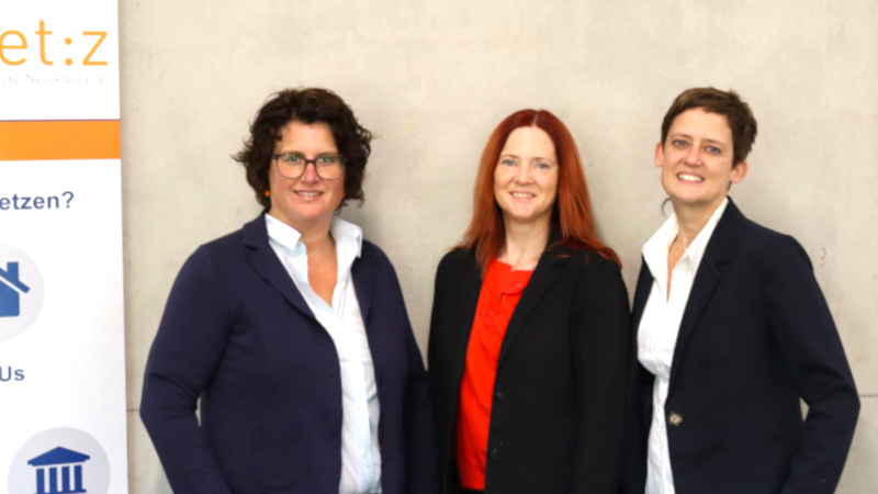 Drei Frauen in Businesskleidung stehen vor einer Betonwand, neben ihnen ist noch ein Teil eines food.net:z-Rollups zu sehen.