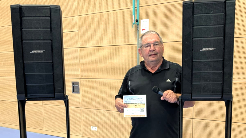 Eine ältere männlich gelesene Person im schwarzen T-Shirt steht zwischen zwei Lautsprecherboxen. In einer Hand hält sie ein Mikrofon, in der anderen eine Förderplakette.