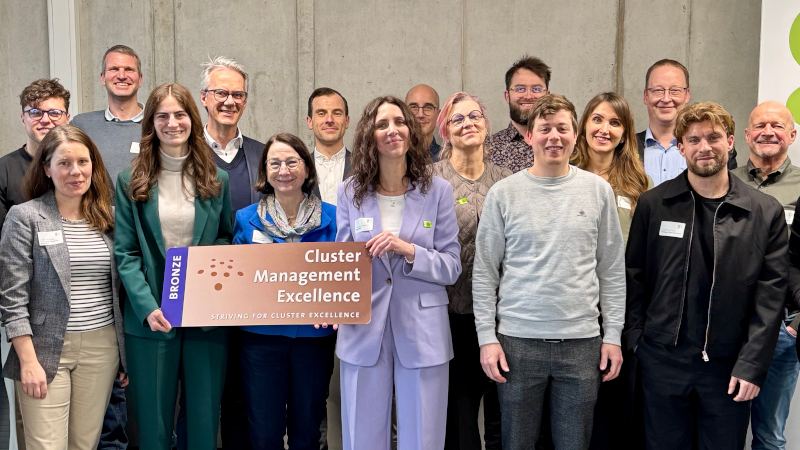 Eine Gruppe von Menschen steht lächelnd vor einer Betonwand. In der vorderen Reihe stehen drei Frauen in Businesskleidung, die gemeinsam ein großes Plakat mit der Aufschrift "Cluster Management Excellence" halten.