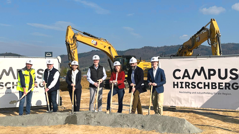 Eine Gruppe von Menschen steht auf einer Baustelle, im Hintergrund sind Bagger zu sehen. Alle sieben Personen haben weiße Baustellenhelme auf. Sie halten je einen Spaten in der Hand und stechen ihn in die Erde.