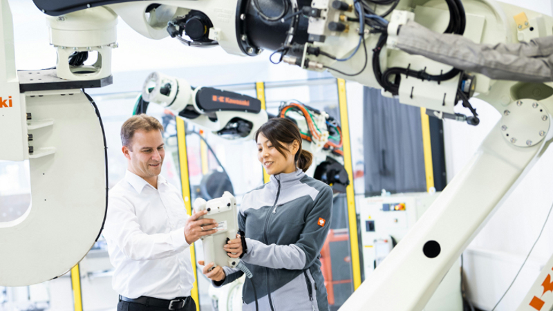 Eine Frau in Arbeitskleidung und ein Mann im Businesshemd stehen zwischen zwei weißen Roboter-Armen und betrachten lächelnd ein weißes Objekt, das die Frau in der Hand hält.