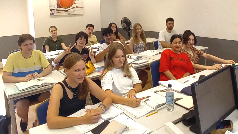 Eine Gruppe Schülerinnen und Schüler sitzt in einem Unterrichtsraum, alle sind sommerlich gekleidet, die meisten lächeln.