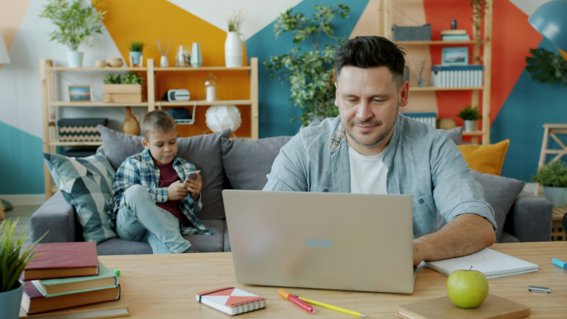 Mann sitzt am Laptop, im Hintergrund sitzt ein Kind auf einem Sofa und schaut auf ein  Smartphone.