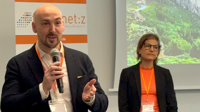 Ein Mann und eine Frau in Businesskleidung. Der Mann steht im Vordergrund, hält ein Mikrofon in der Hand und spricht. 