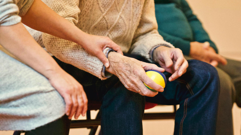 Bildausschnitt: Drei Menschen sitzen in einer Reihe, man sieht nur die Beine knapp unterhalb der Knie und einen Teil des Torsos. Rechts sitzen zwei alte Männer, links eine junge Frau. Sie legt eine Hand auf das Handgelenk des Mannes in der Mitte.